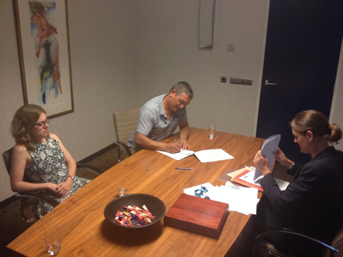 Foto van 3 mensen om een tafel waarbij 1 iemand papieren ondertekend