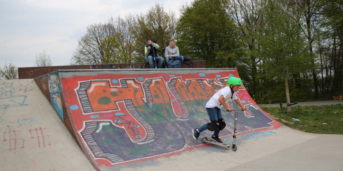 Een jonge stuntstepper met groene helm in actie op de skatebaan; twee jongens kijken toe