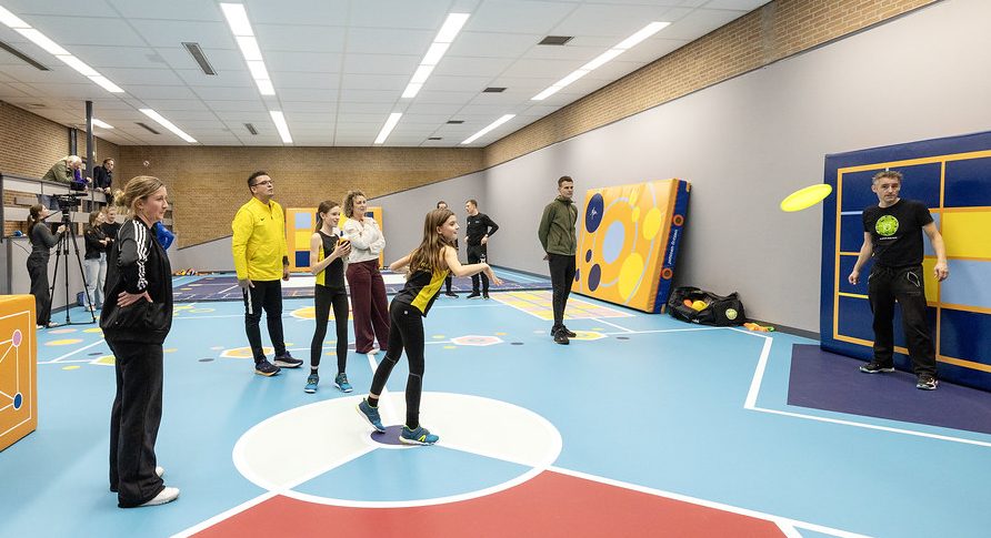 Kind gooit frisbee in nieuwe ASM-sportzaal. Omstanders kijken toe.