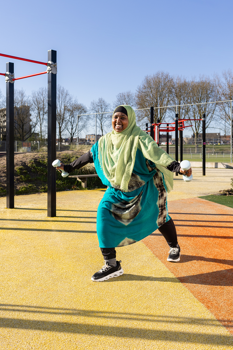 Een vrolijke vrouw met hoofddoek doet oefeningen met gewichtjes op een openbare beweegplek