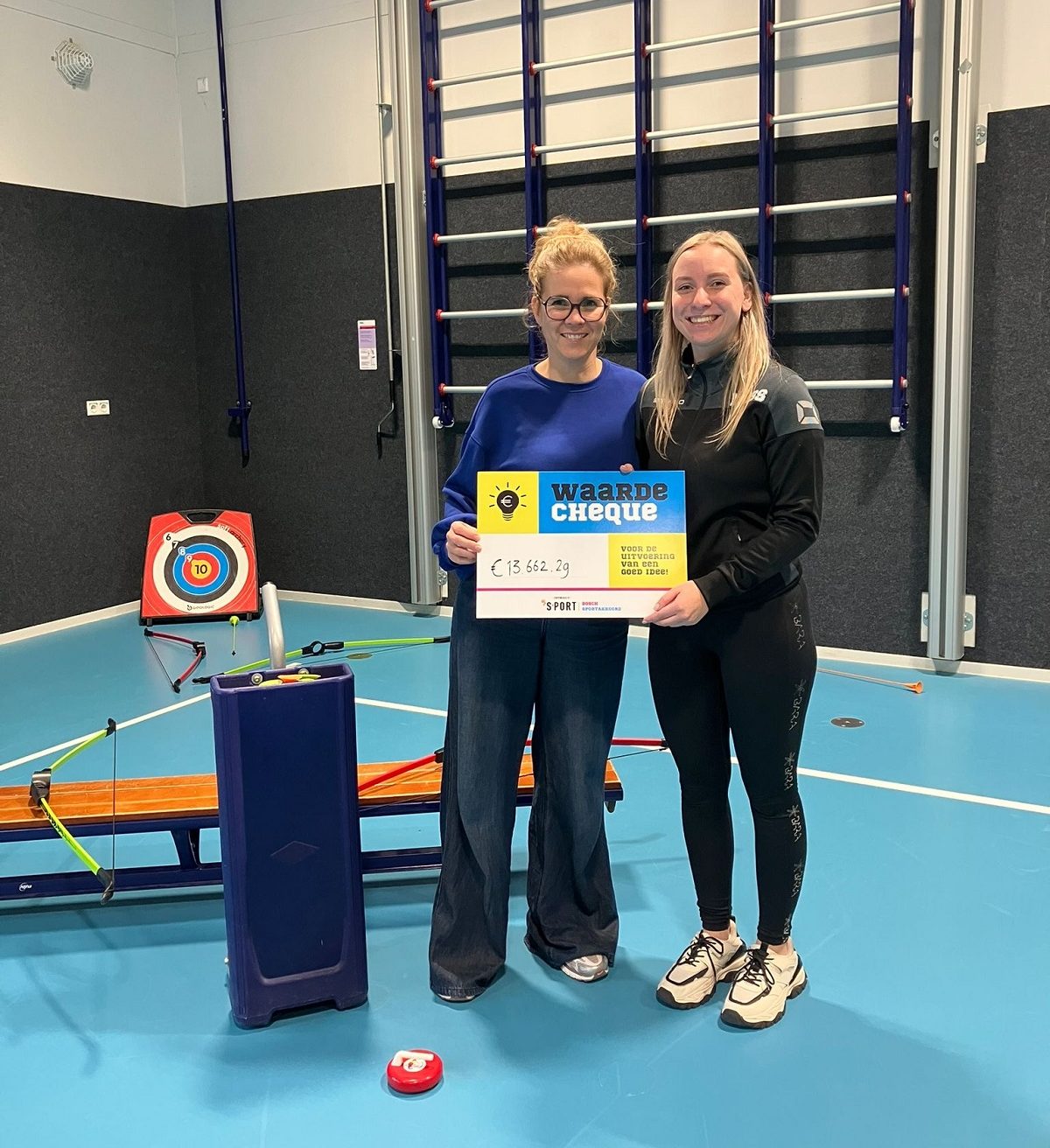 Twee vertegenwoordigers van HUB Rosmalen ontvangen in gymzaal een stimuleringscheque van Bosch Sportakkoord voor nieuw beweegproject; op achtergrond allerlei spelmaterialen