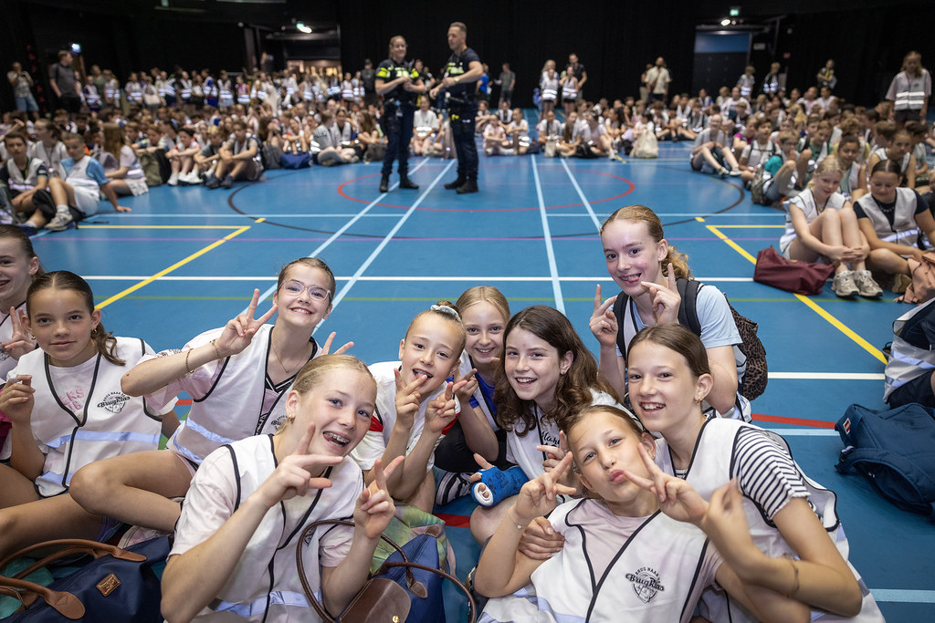 Meisjes op voorgrond die met hun handen een V-teken maken in een sportzaal; daarachter veel andere kinderen