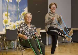Twee vrouwen voeren strekoefening uit met elastieken; de een zittend, de ander staand