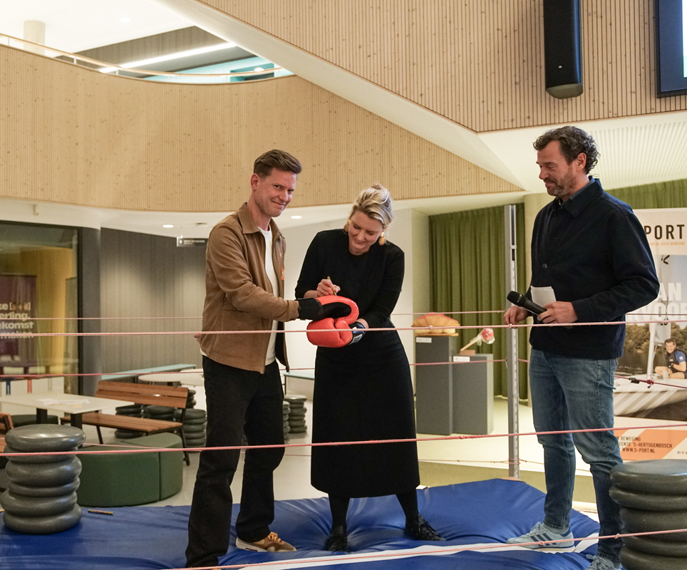 Mede-initiatiefnemer en voorzitter Michiel van Rijn van Alkemade en sport-wethouder Marianne van der Sloot zetten handtekeningen op een paar bokshandschoenen; dagvoorzitter kijkt toe