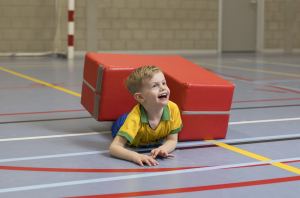 Een lachend jongetje ligt in een sportzaal bij en halfonder een rood zacht blok