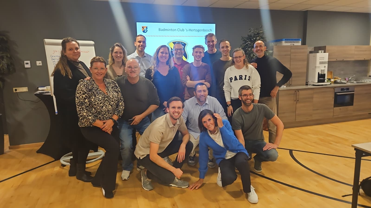 Een groep mensen met daarachter een scherm waarop de tekst 'Badminton Club 's-Hertogenbosch te zien is'