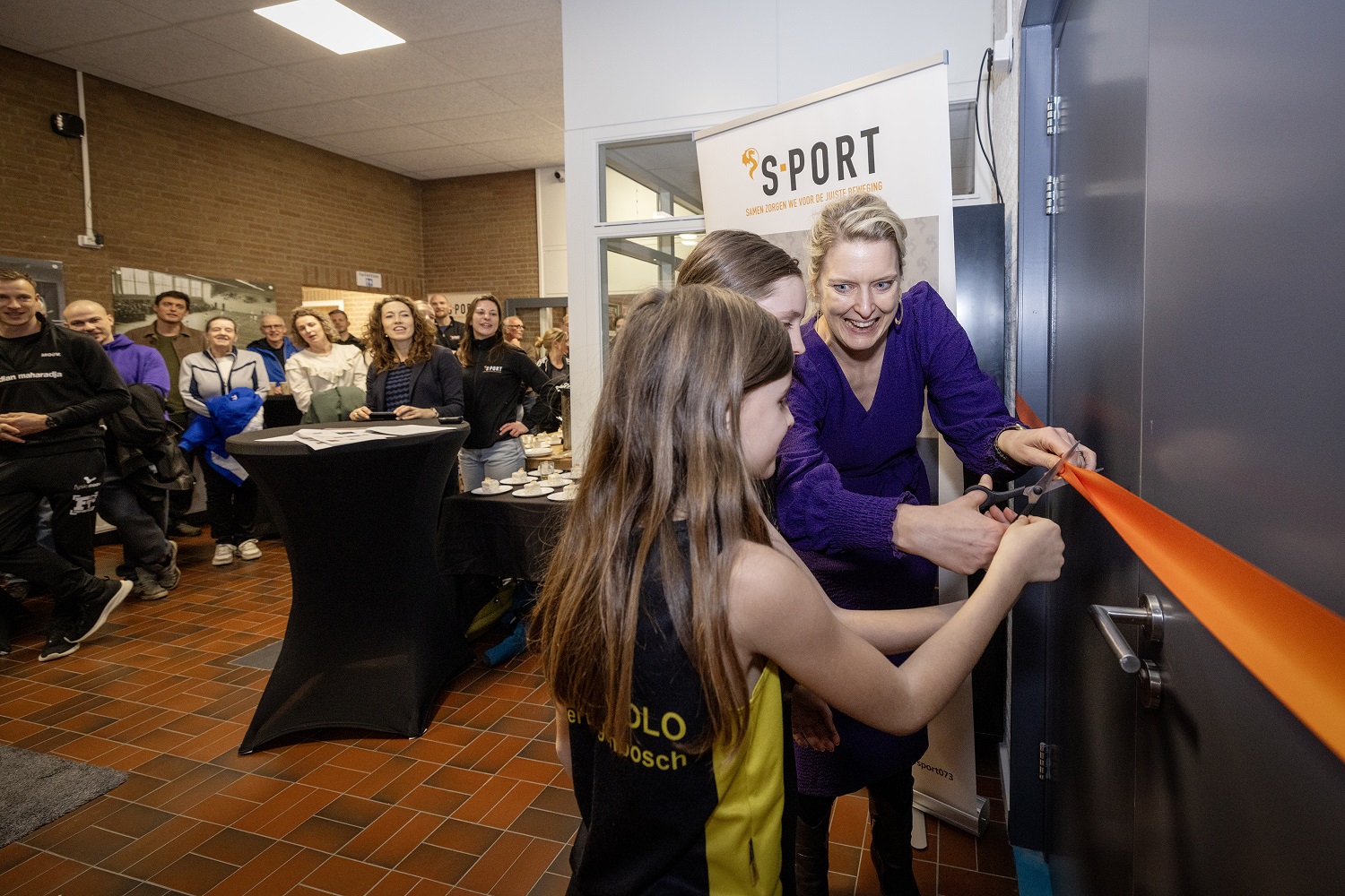 Wethouder Marianne van der Sloot knipt samen met twee meisjes oranje lint voor deur nieuwe sportzaal door