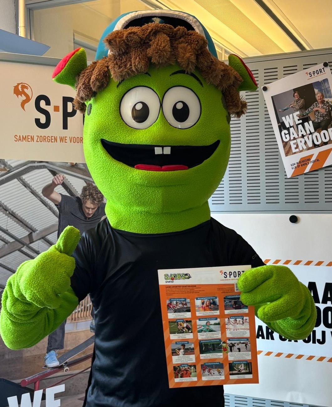 De groene mascotte Sjors Sportief geeft een duim omhoog en houdt een flyer omhoog voor een ´S-PORT‑promotiewand