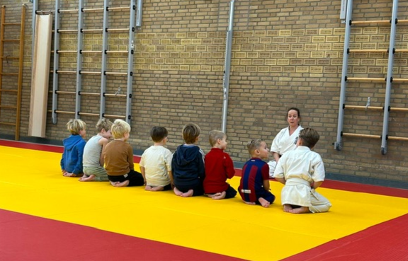 Acht kinderen op de rug gezien zittend op een mat, voor hen de karate-instructrice