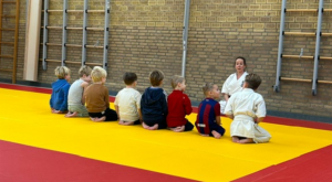 Acht kinderen op de rug gezien zittend op een mat, voor hen de karate-instructrice