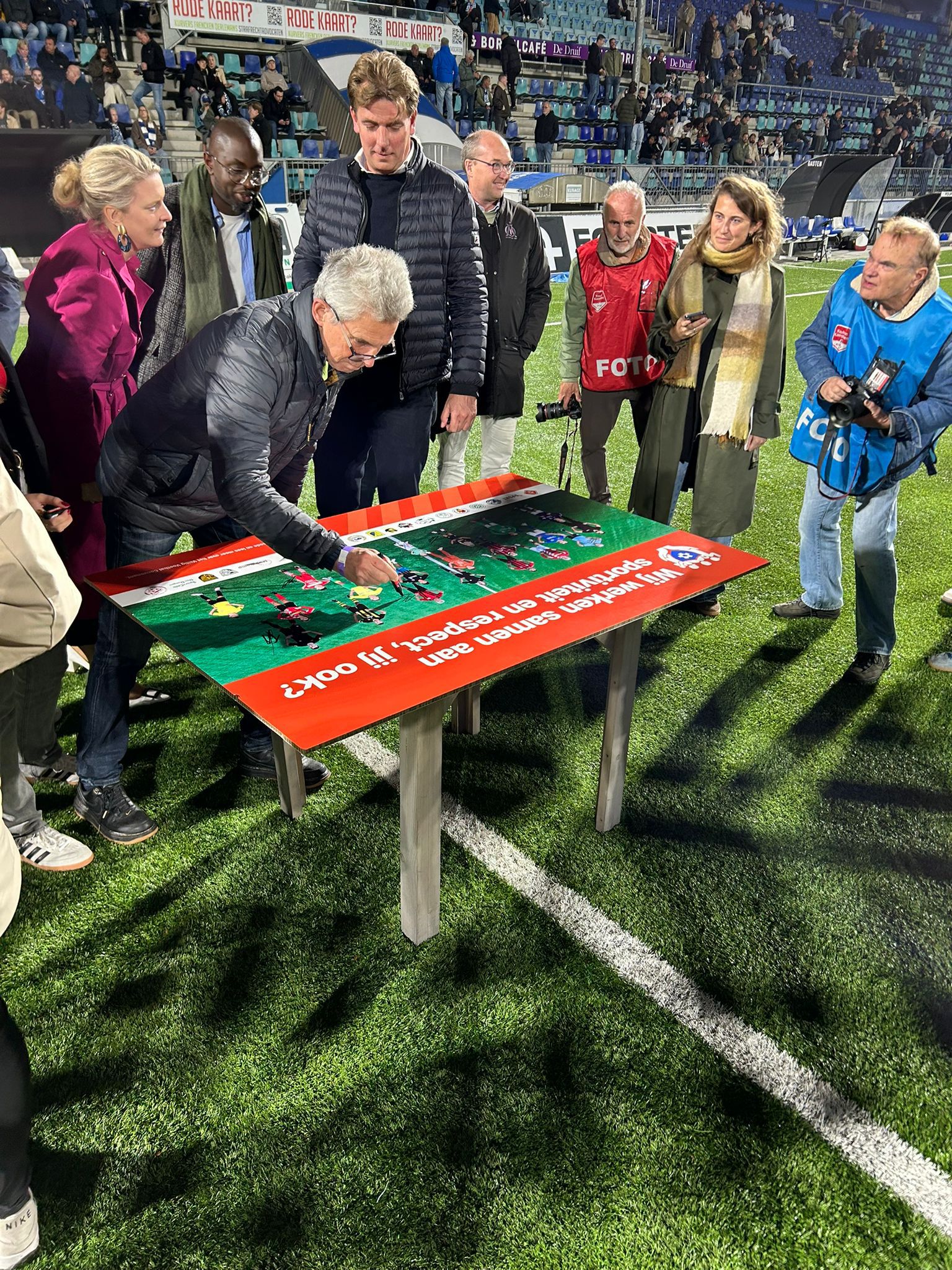 Menens ondertekenen Veilig Voetbal Convenant in stadion, wethouder en fotografen kijken toe