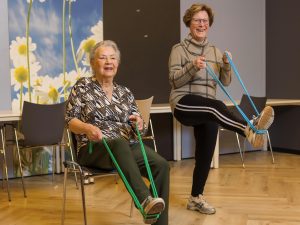 Gerda (91) en Marij (78) bewegen zich fit: Gerda zit in een stoel, Marij staat lachend naast haar