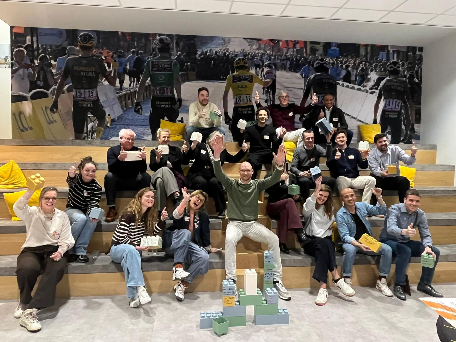 Een groep mensen steekt handen of duimen in de lucht, zittend op tredes met een grote sportieve foto op de achtergrond; voor hen staan grote legoblokken